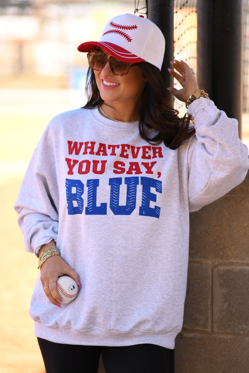 Woman wearing ash gray “Whatever You Say Blue” baseball graphic sweatshirt with red and blue lettering, styled with a baseball cap and holding a baseball at a ball field.