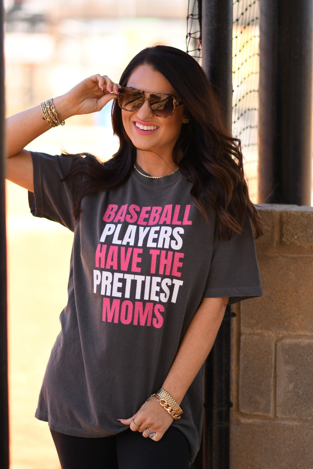Woman wearing a pepper Comfort Colors graphic tee that says "Baseball Players Have The Prettiest Moms" in pink and white lettering, styled with sunglasses and black leggings at a baseball field.