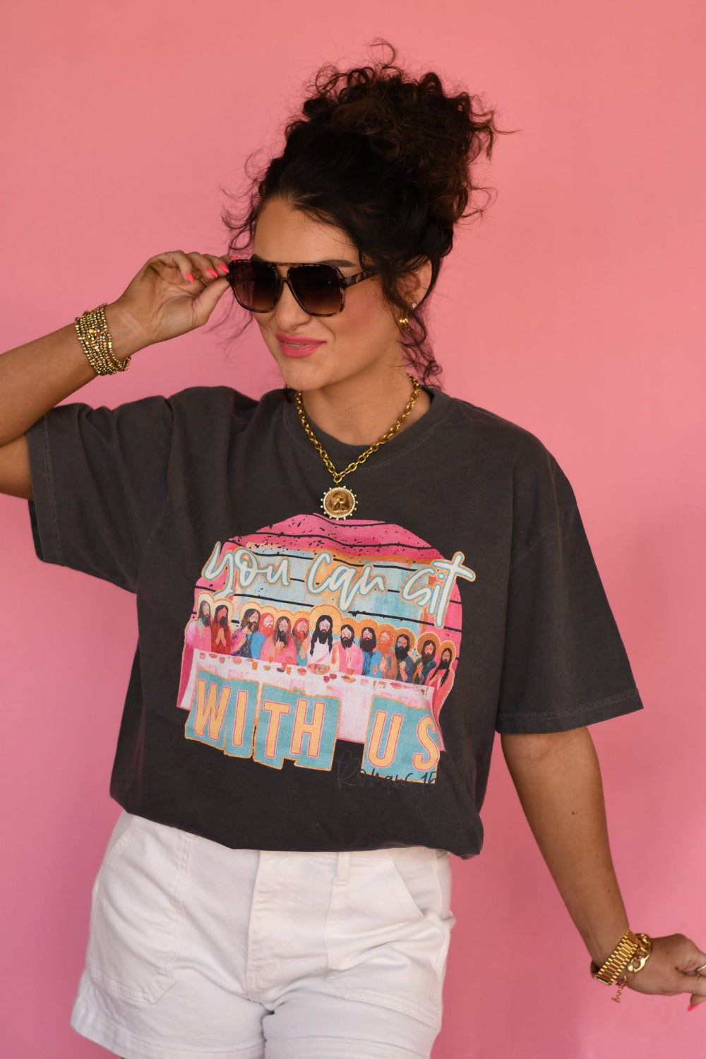 Woman wearing a Pepper Comfort Colors “You Can Sit With Us” Christian graphic tee with faith-based design, styled with white shorts against a pink background.