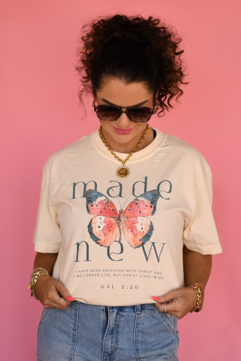 Woman wearing ivory Made New butterfly Christian graphic tee with Galatians 2:20 scripture, styled with denim shorts and gold jewelry against pink background