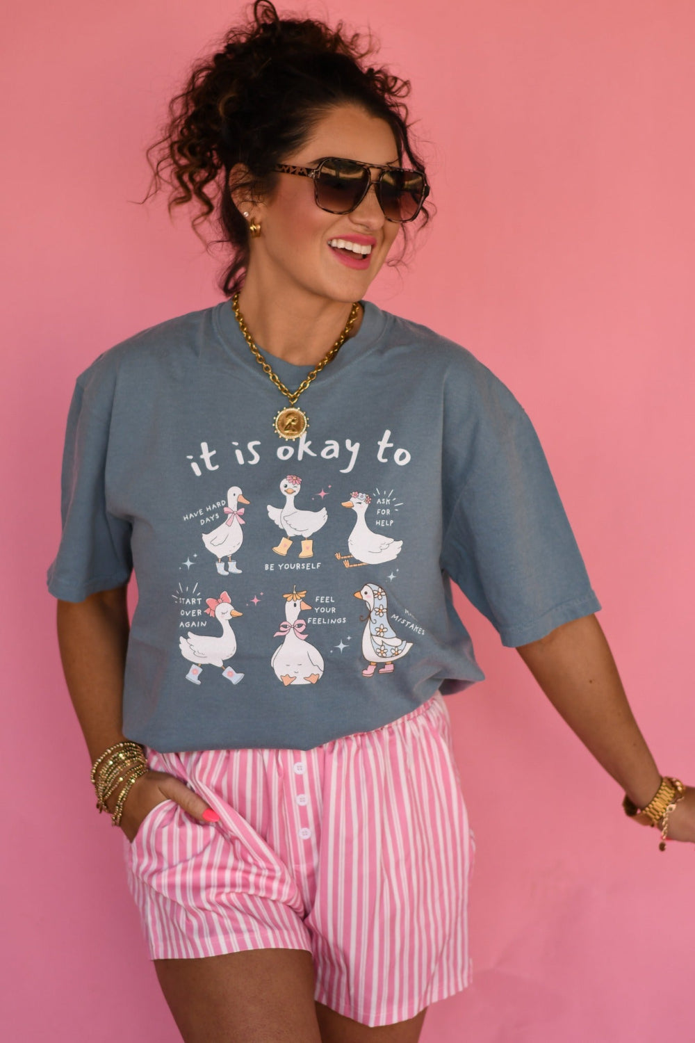 Woman wearing Ice Blue Comfort Colors “It Is Okay to Geese” graphic tee featuring cute illustrated geese with positive sayings, styled with pink striped shorts against a pink background.