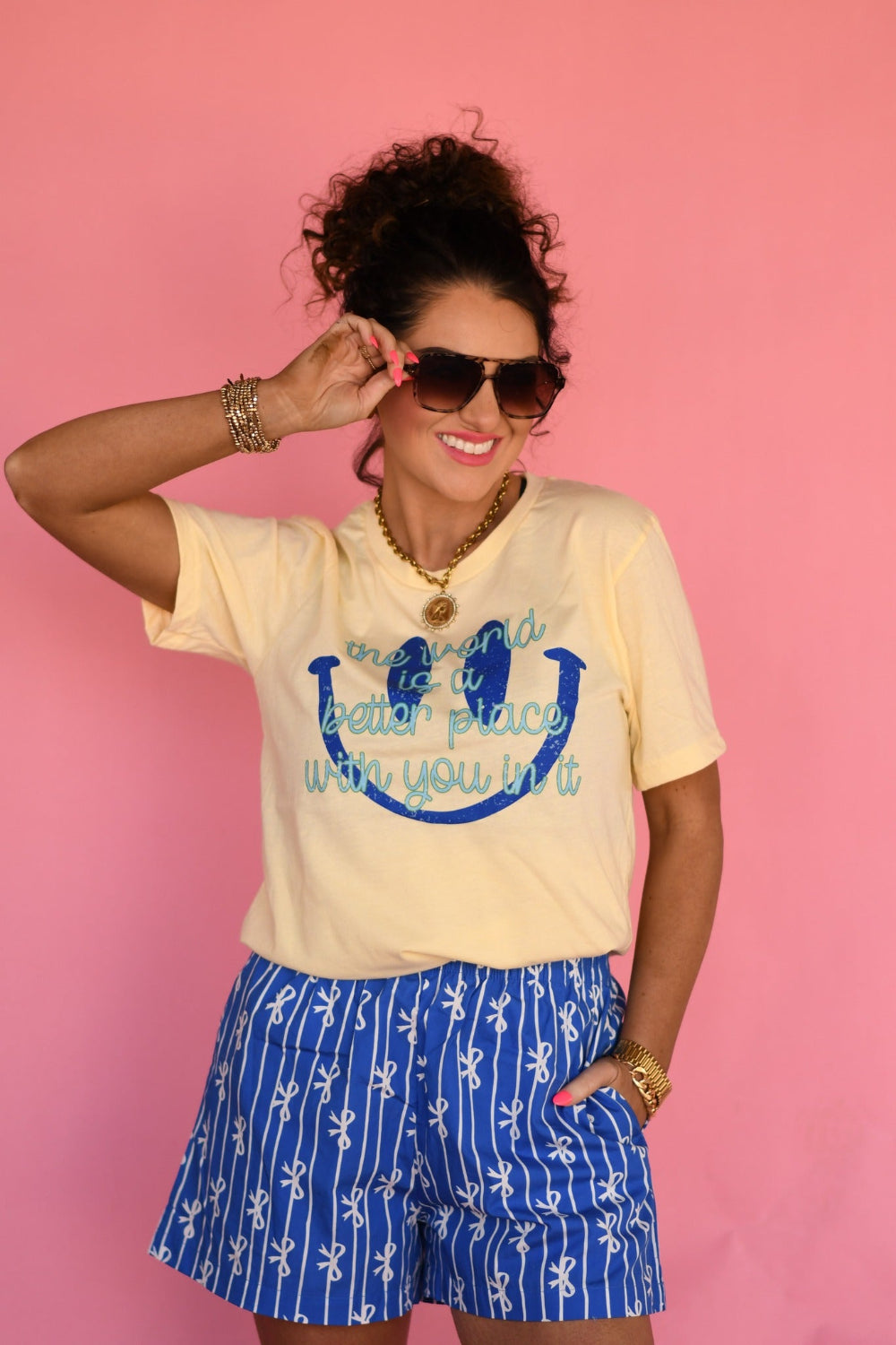 Woman wearing a butter yellow “The World Is a Better Place With You In It” graphic Bella Canvas tee styled with blue patterned shorts against a pink background.
