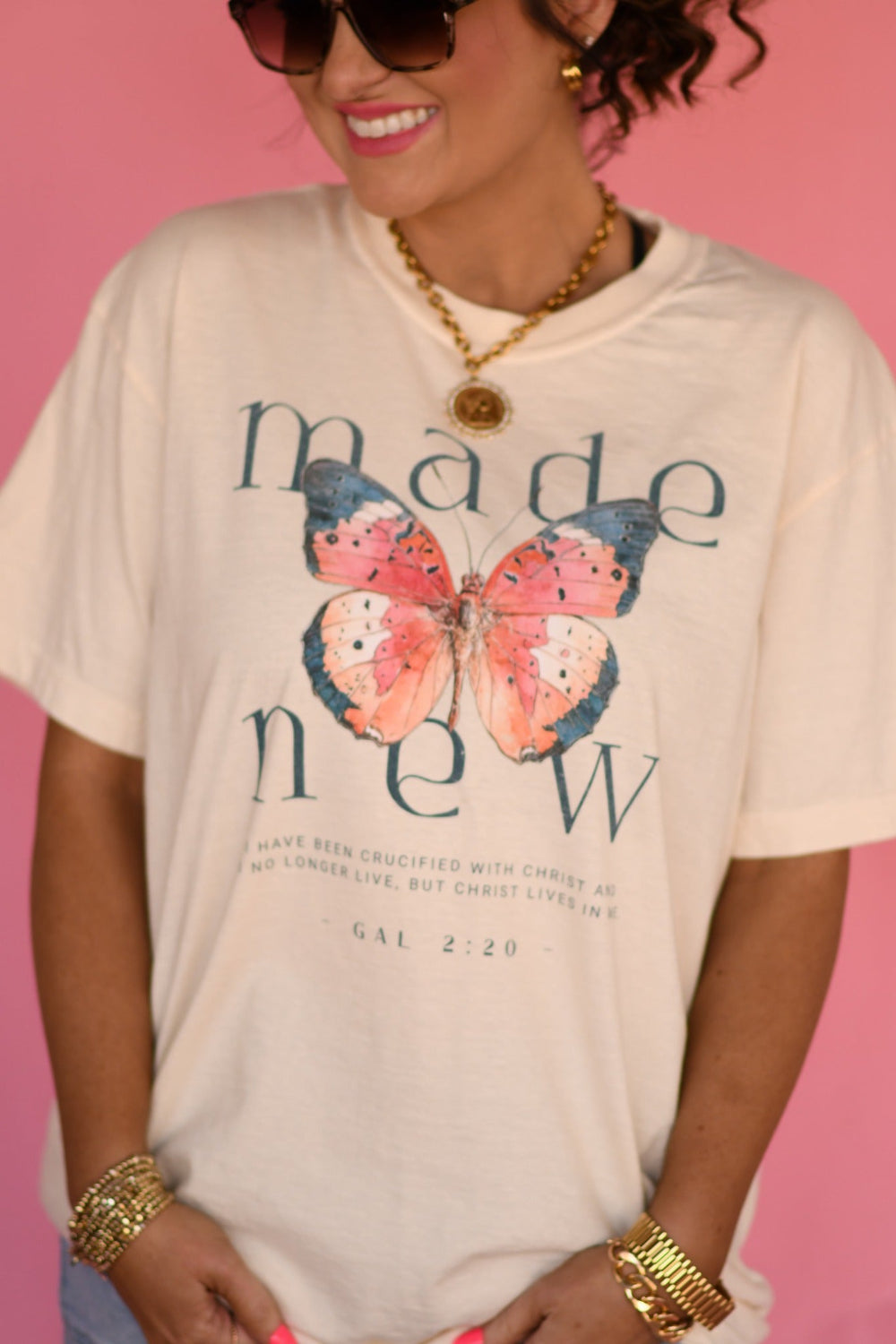 Woman wearing ivory Made New butterfly Christian graphic tee with Galatians 2:20 scripture, styled with denim shorts and gold jewelry against pink background