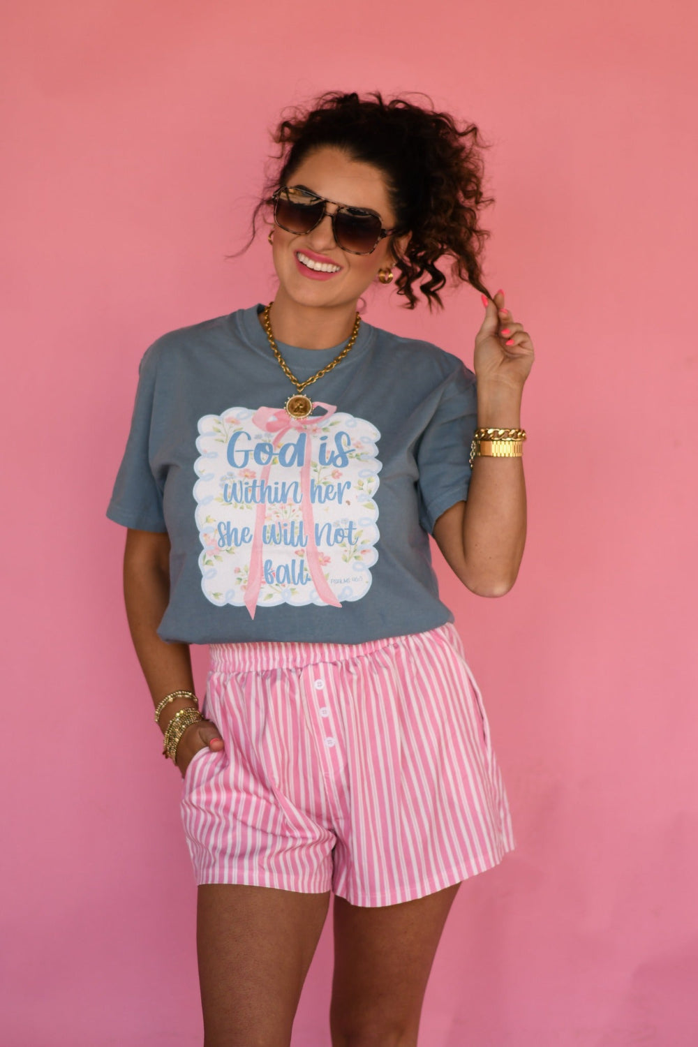 Woman wearing Ice Blue Comfort Colors “God Is Within Her She Will Not Fall” Christian graphic tee with pink bow floral design from Psalm 46:5