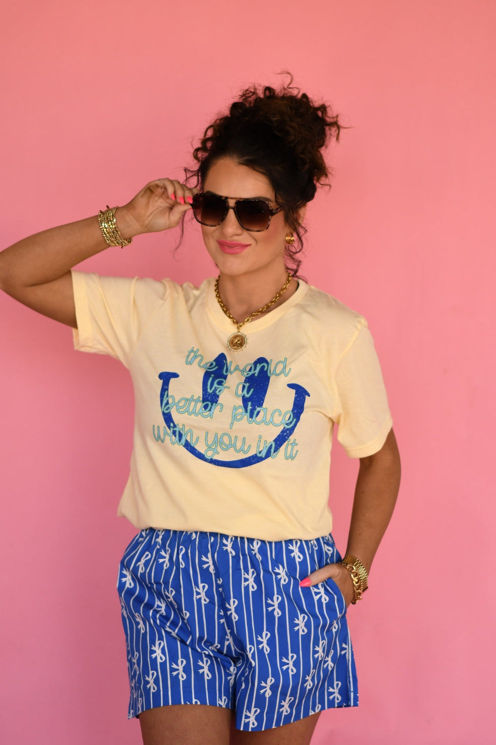 Woman wearing a butter yellow “The World Is a Better Place With You In It” graphic Bella Canvas tee styled with blue patterned shorts against a pink background.