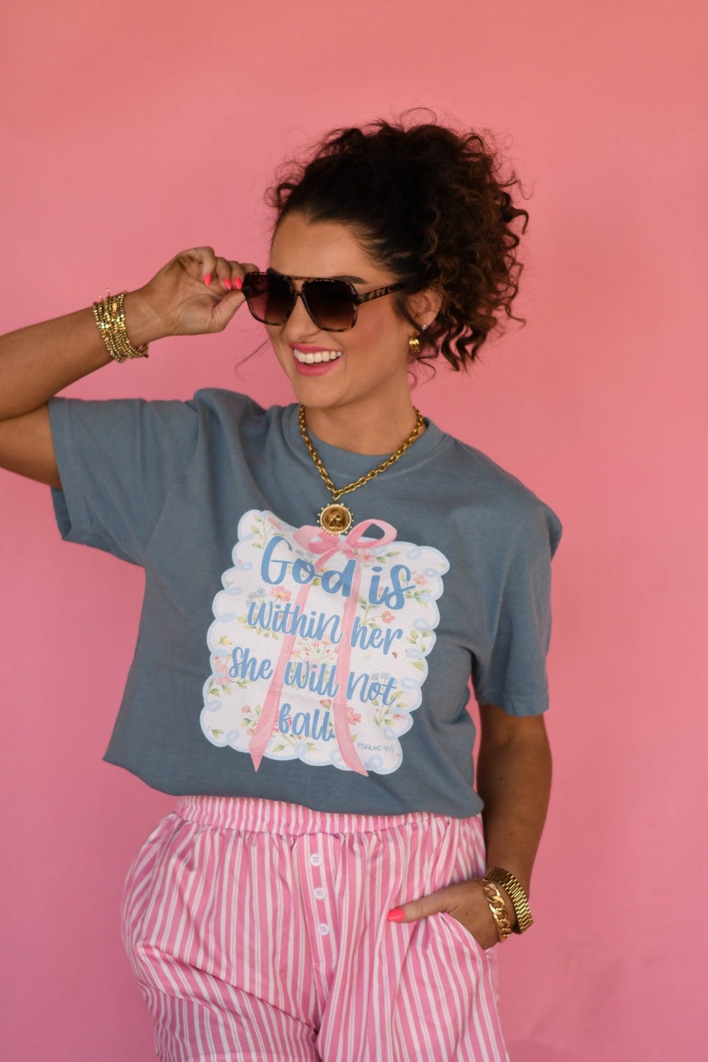 Woman wearing Ice Blue Comfort Colors “God Is Within Her She Will Not Fall” Christian graphic tee with pink bow floral design from Psalm 46:5