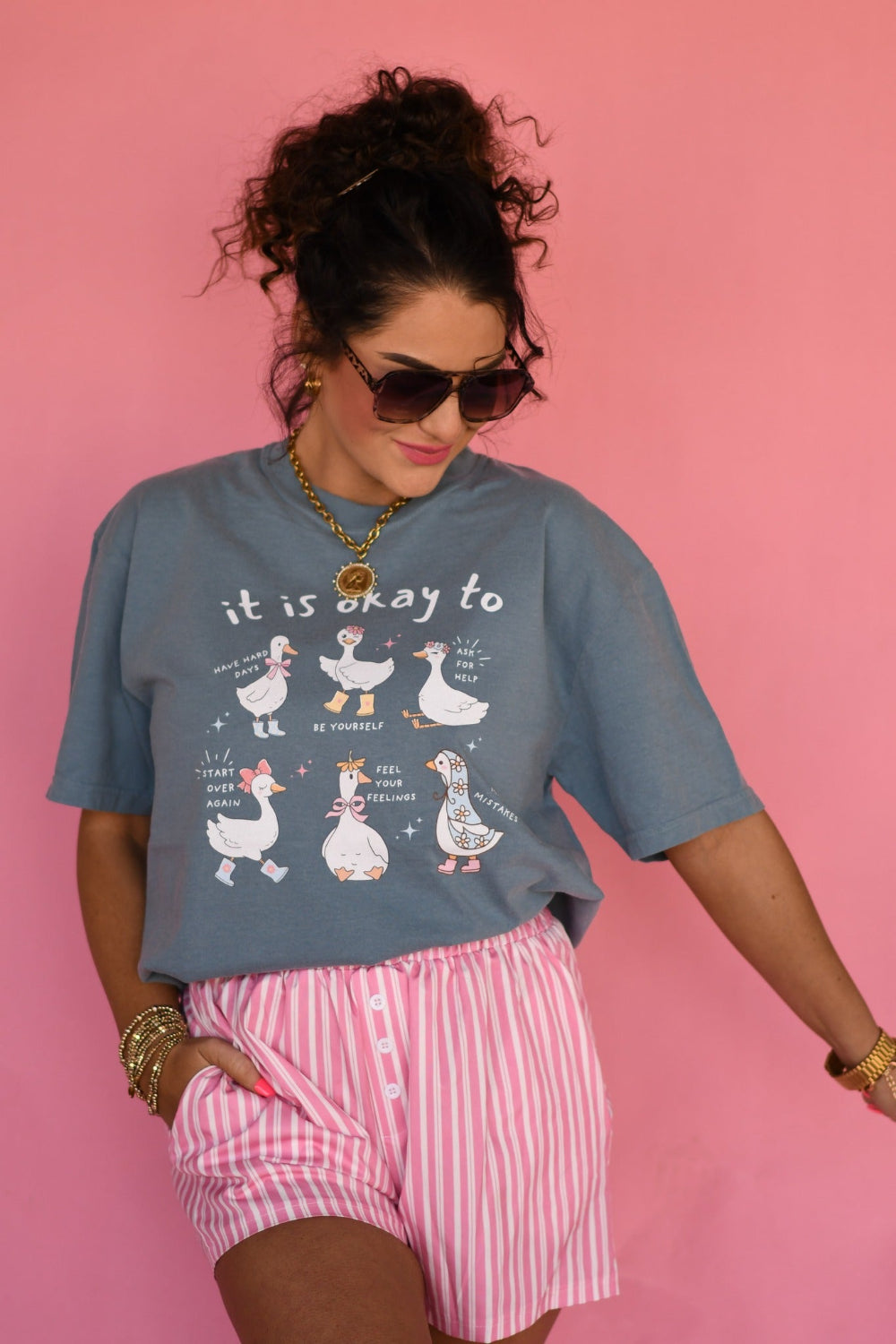 Woman wearing Ice Blue Comfort Colors “It Is Okay to Geese” graphic tee featuring cute illustrated geese with positive sayings, styled with pink striped shorts against a pink background.