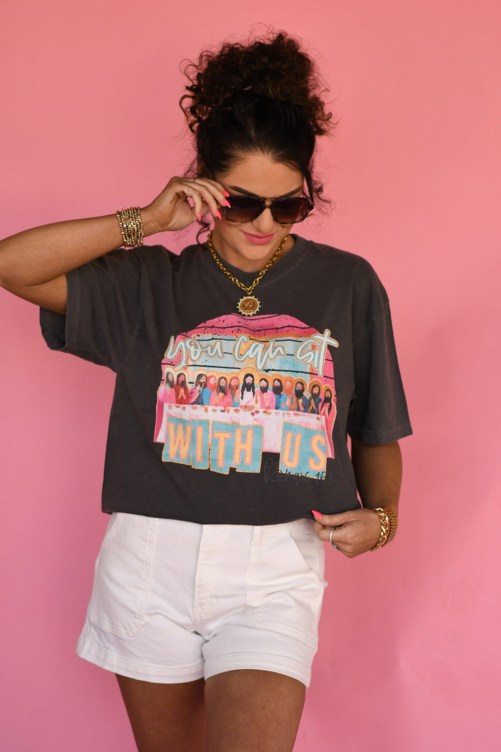 Woman wearing a Pepper Comfort Colors “You Can Sit With Us” Christian graphic tee with faith-based design, styled with white shorts against a pink background.
