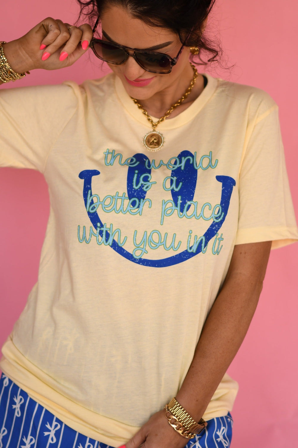 Woman wearing a butter yellow “The World Is a Better Place With You In It” graphic Bella Canvas tee styled with blue patterned shorts against a pink background.