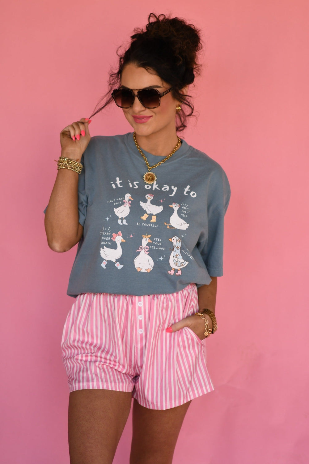 Woman wearing Ice Blue Comfort Colors “It Is Okay to Geese” graphic tee featuring cute illustrated geese with positive sayings, styled with pink striped shorts against a pink background.