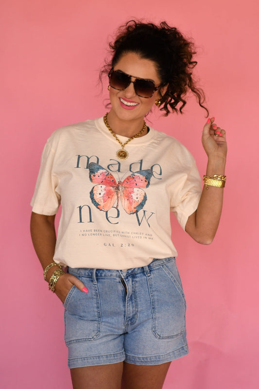 Woman wearing ivory Made New butterfly Christian graphic tee with Galatians 2:20 scripture, styled with denim shorts and gold jewelry against pink background