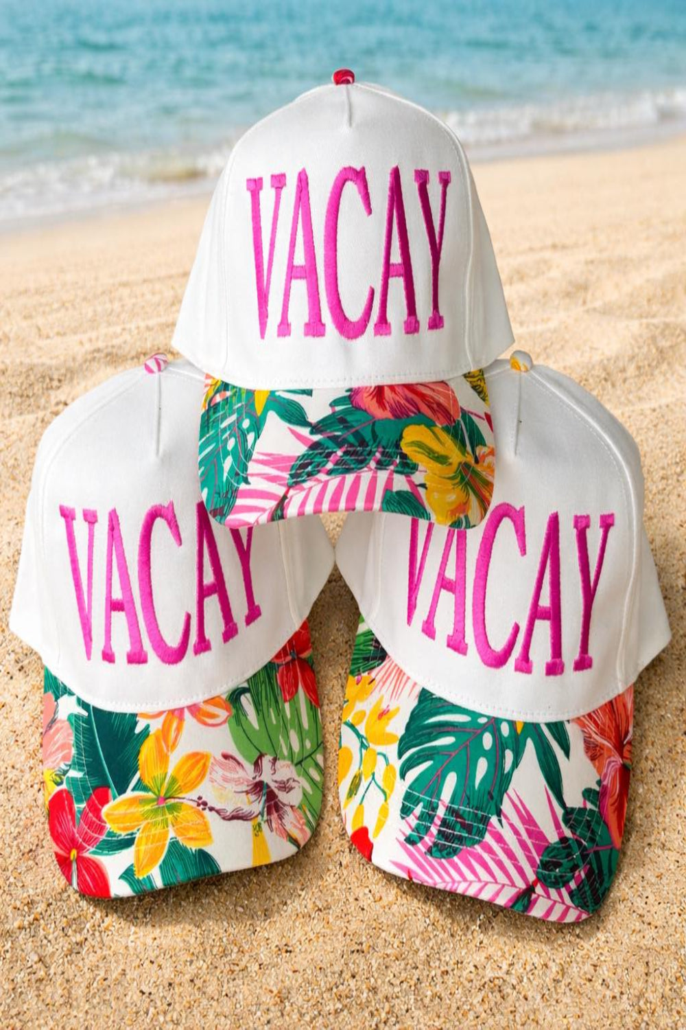 White trucker hat with pink VACAY embroidery and tropical floral brim displayed on beach sand