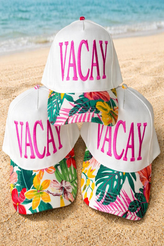 White trucker hat with pink VACAY embroidery and tropical floral brim displayed on beach sand