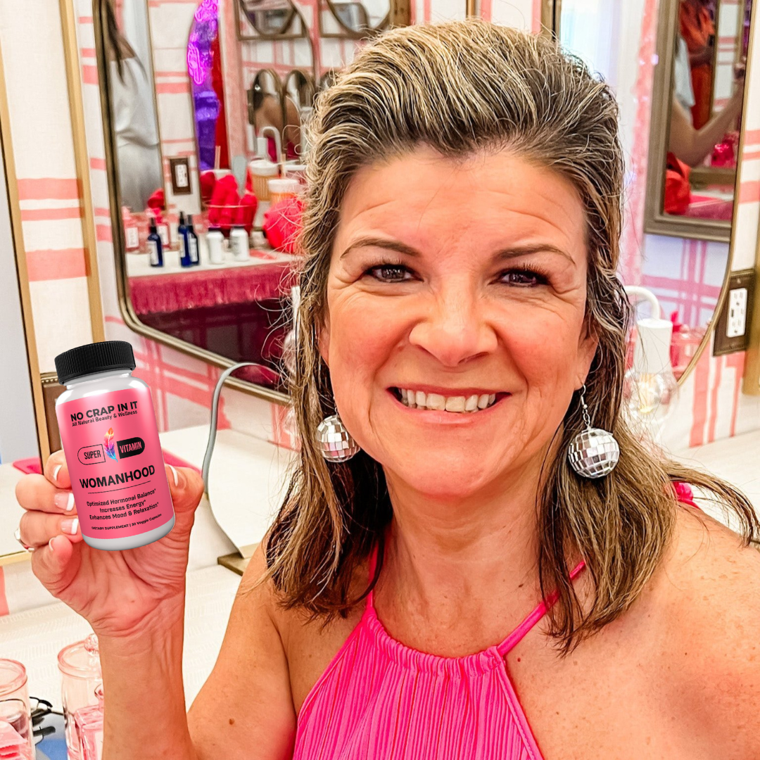 A bottle of 'No Crap In It' Womanhood Super Vitamin Supplement with pink label, black cap, and text indicating optimized hormonal balance, increased energy, and enhanced mood and relaxation.