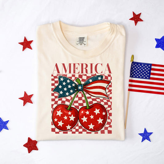 Cream graphic tee featuring red “America” text, patriotic cherries with stars, an American flag bow, and red checkerboard background.
