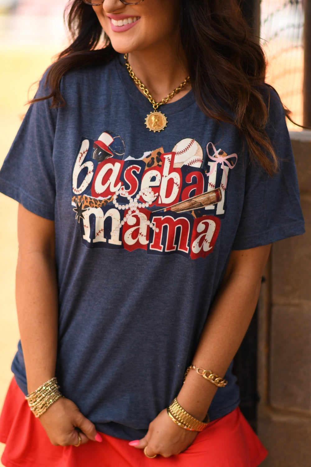 Baseball Mama Icons graphic tee in heather navy worn by woman with red athletic skirt at baseball field