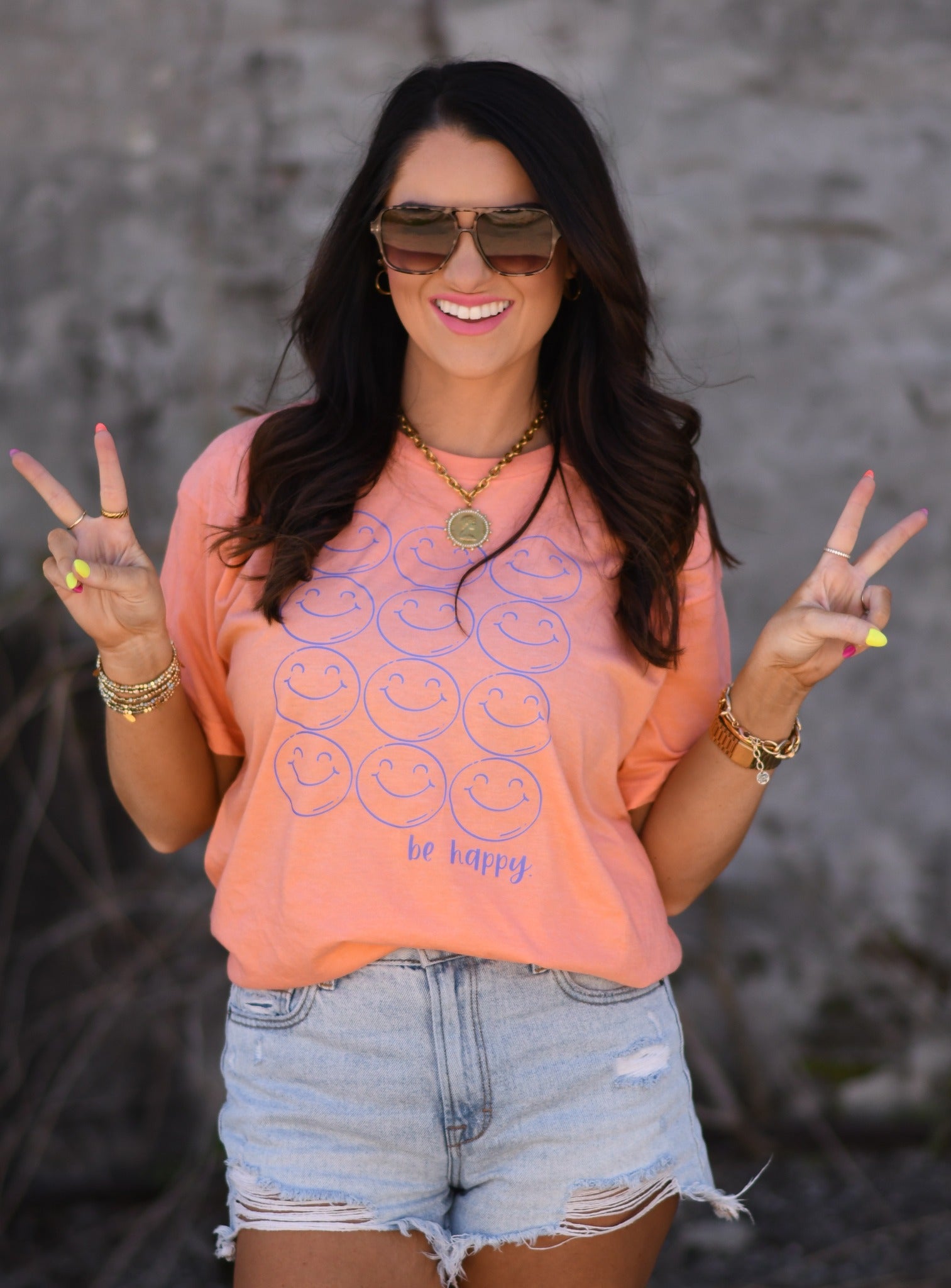 Woman wearing a heather cantaloupe Be Happy smiley face graphic tee with denim shorts and sunglasses, holding peace signs