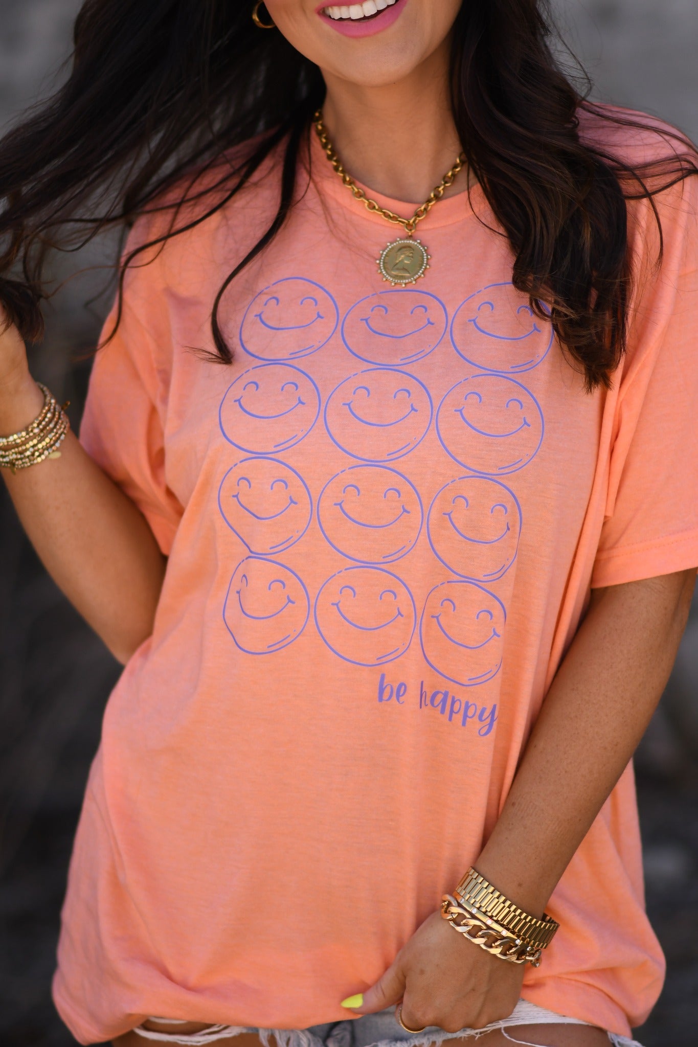 Woman wearing a heather cantaloupe Be Happy smiley face graphic tee with denim shorts and sunglasses, holding peace signs
