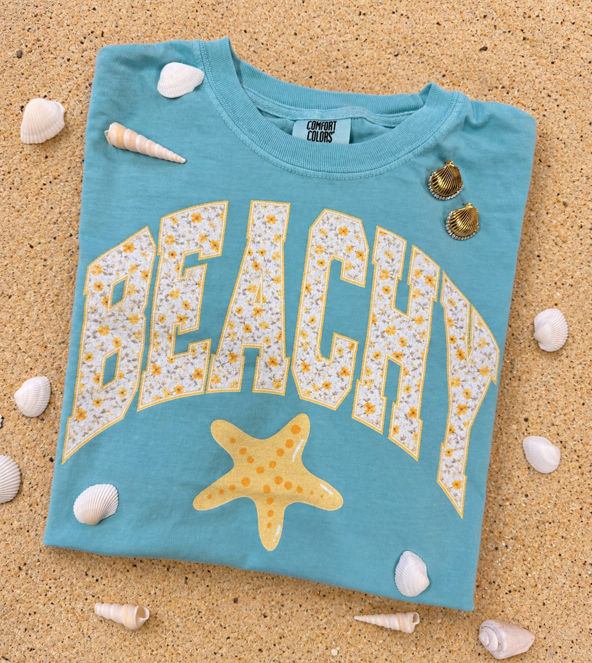 Blue Comfort Colors beachy graphic tee with floral varsity lettering and yellow starfish design styled on sand with seashells.