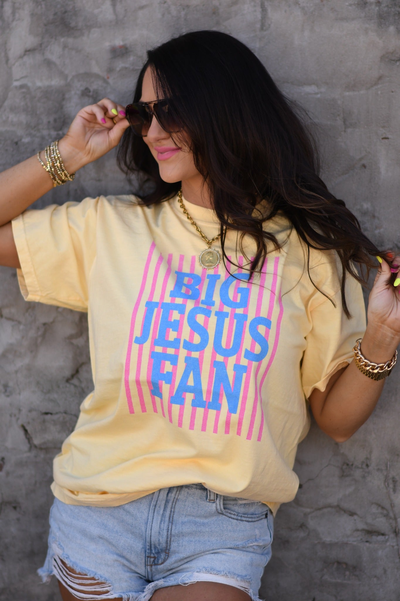 Woman wearing butter yellow Big Jesus Fan striped graphic tee with pink and blue design, styled with denim shorts and sunglasses