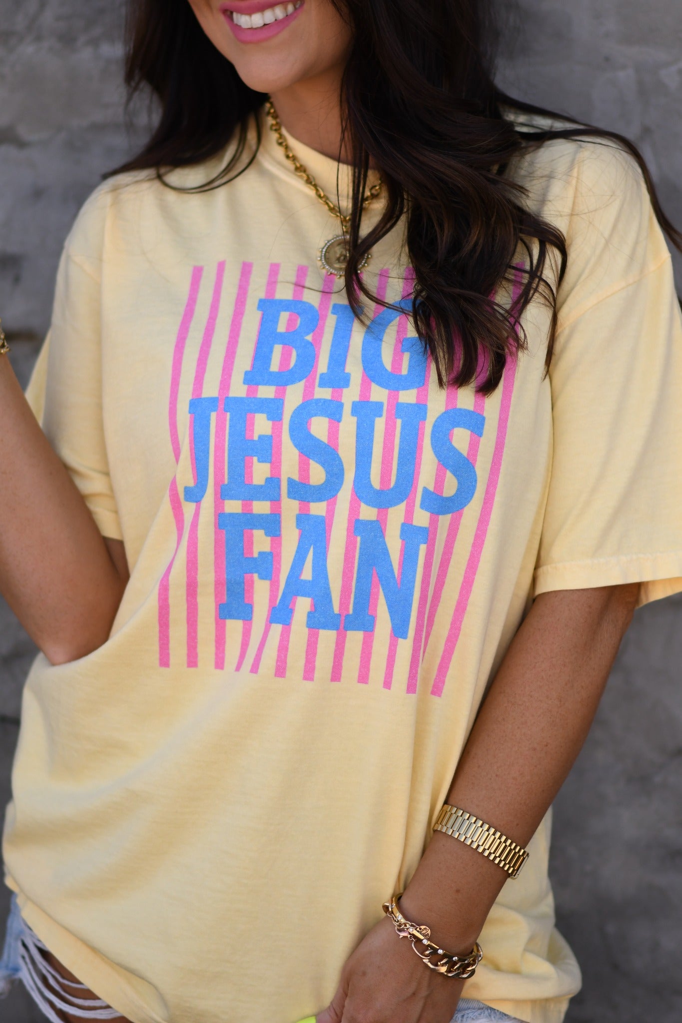 Woman wearing butter yellow Big Jesus Fan striped graphic tee with pink and blue design, styled with denim shorts and sunglasses