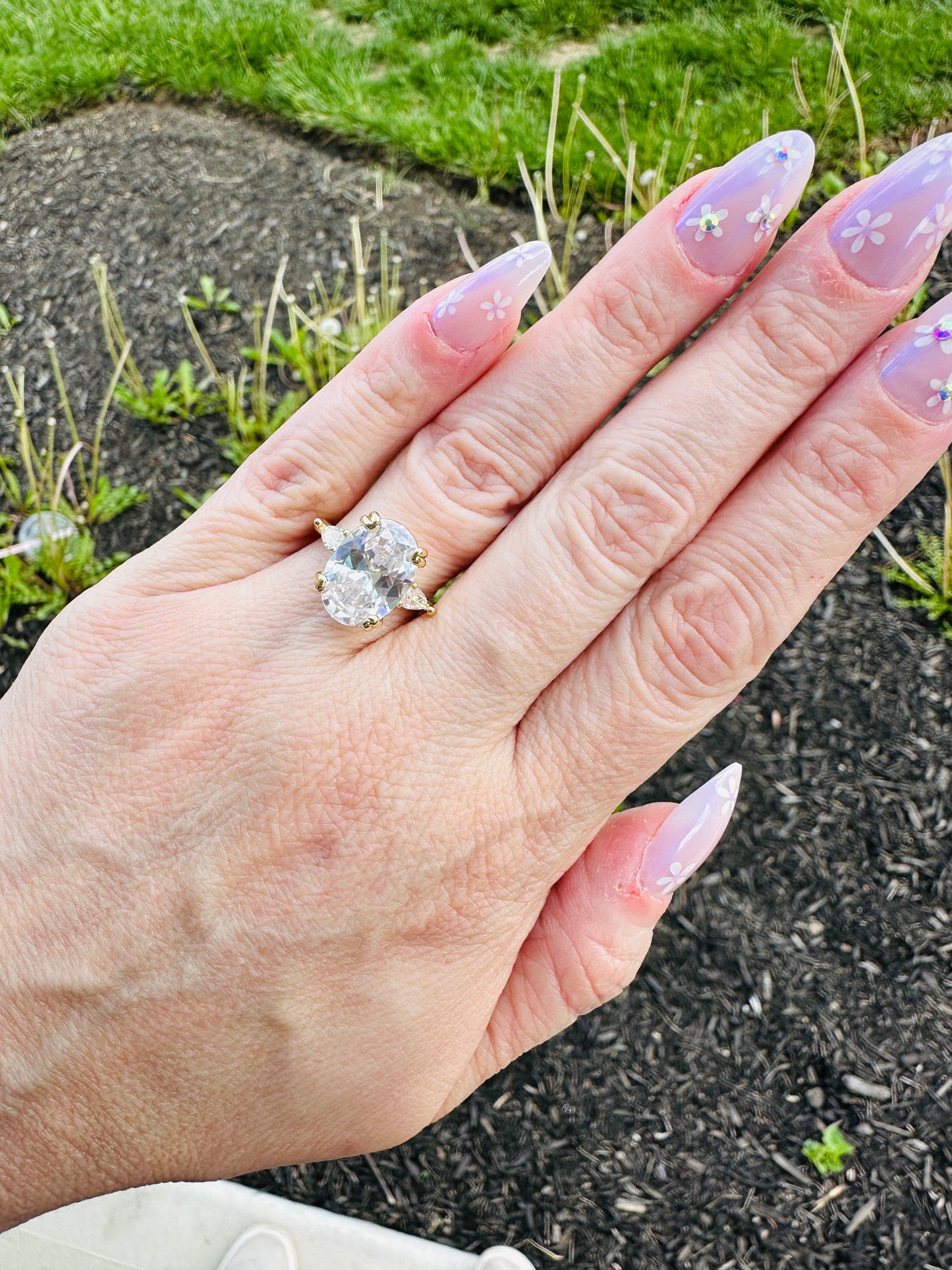 Hand wearing Liora Luxe oval three-stone gold plated ring with large cubic zirconia center stone and floral nail art