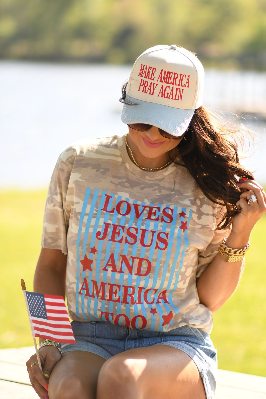 Woman wearing a natural camo graphic tee that says "Loves Jesus and America Too" with a patriotic hat and American flag outdoors