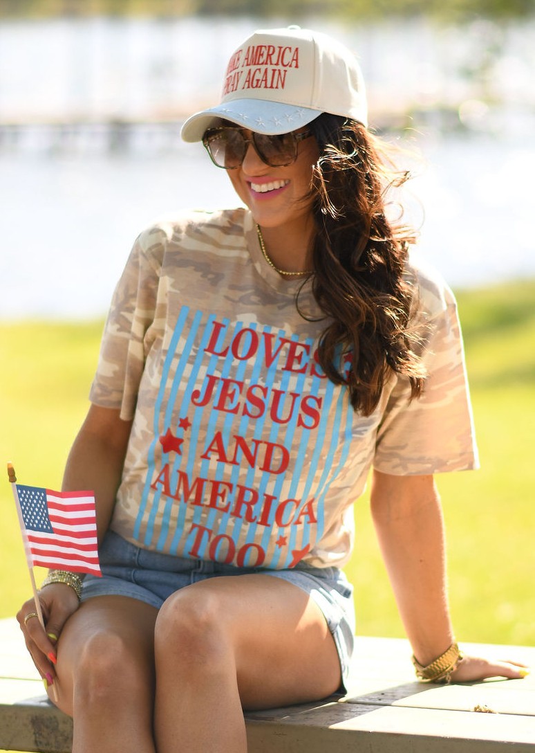 Woman wearing a natural camo graphic tee that says "Loves Jesus and America Too" with a patriotic hat and American flag outdoors