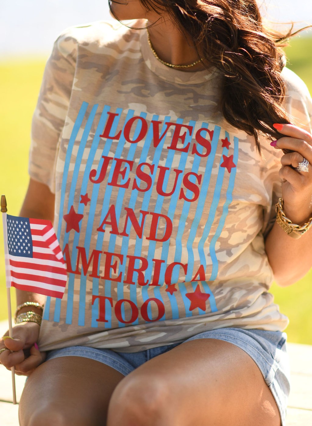 Woman wearing a natural camo graphic tee that says "Loves Jesus and America Too" with a patriotic hat and American flag outdoors