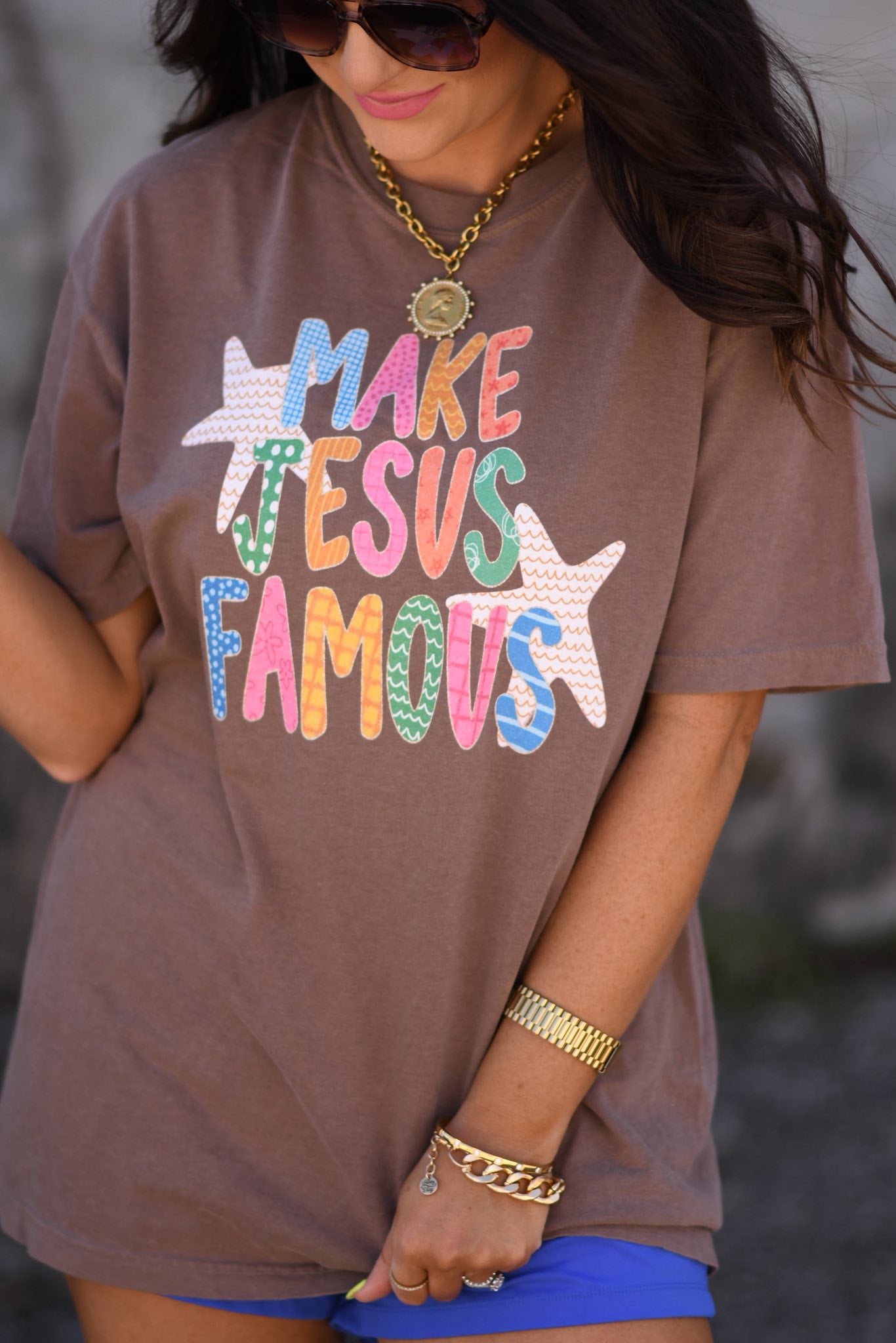 Woman wearing espresso brown “Make Jesus Famous” colorful graphic t-shirt with sunglasses and gold jewelry, styled with blue shorts for a summer outfit