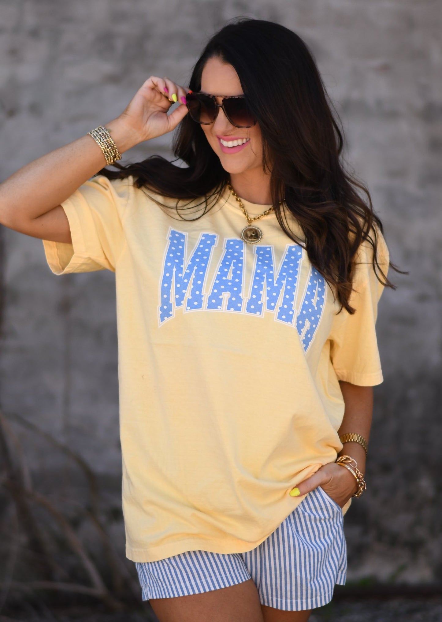 Woman wearing butter yellow MAMA graphic tee with blue polka dots paired with striped shorts and sunglasses