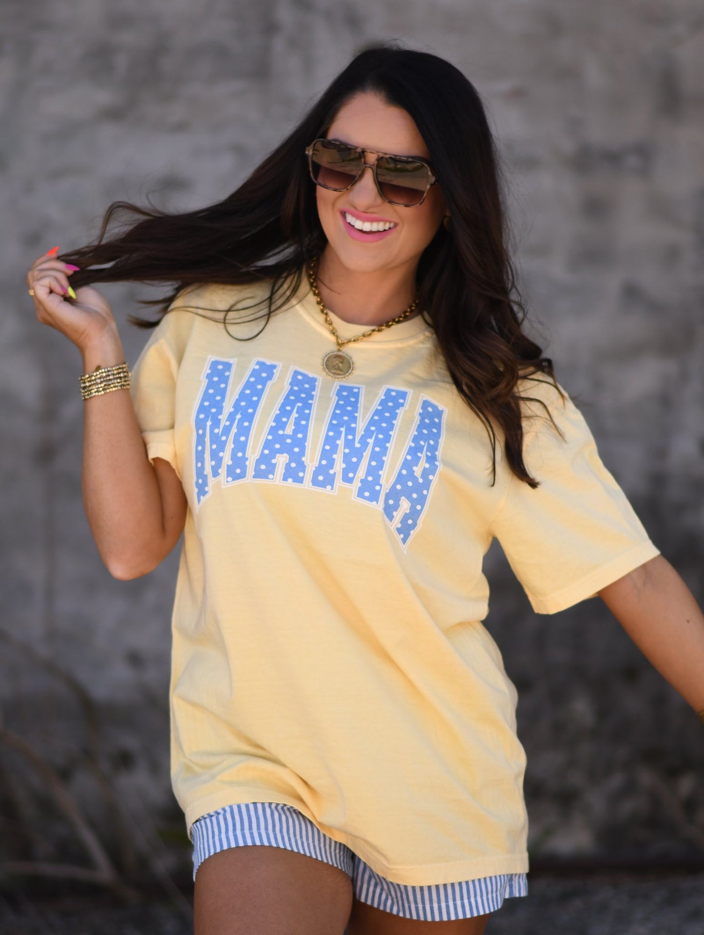 Woman wearing butter yellow MAMA graphic tee with blue polka dots paired with striped shorts and sunglasses