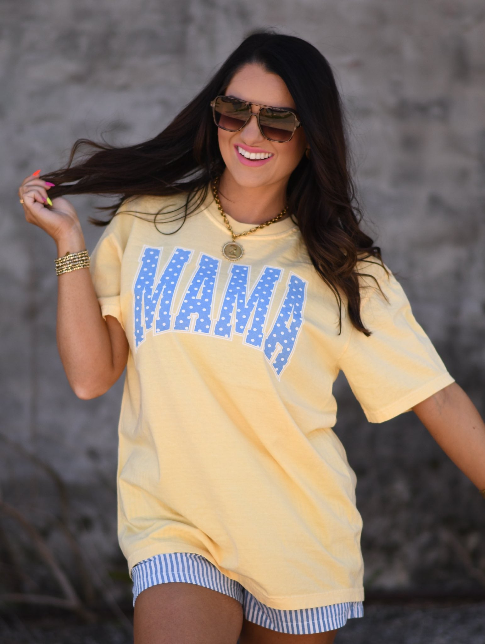 Woman wearing butter yellow MAMA graphic tee with blue polka dots paired with striped shorts and sunglasses