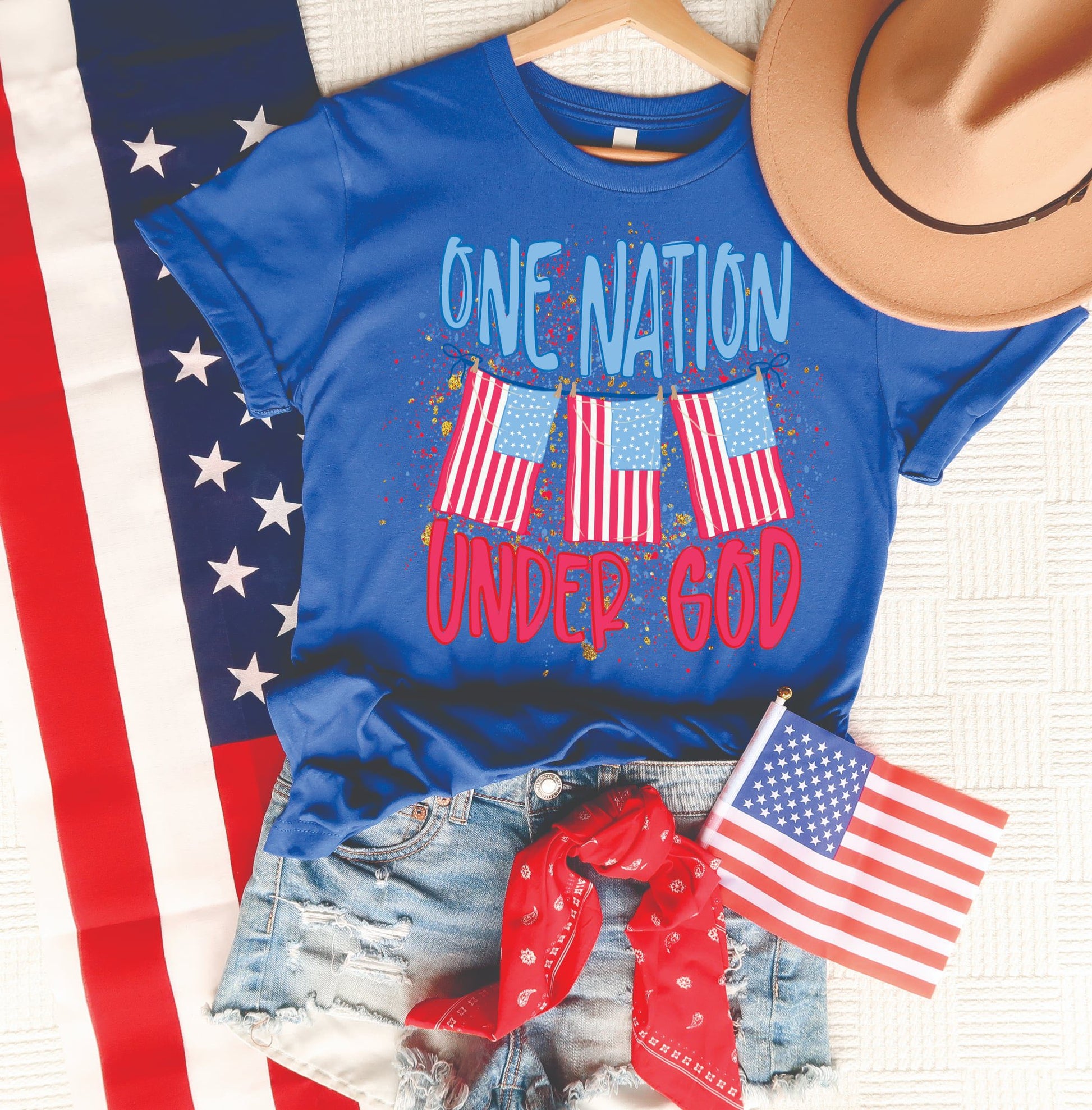 One Nation Under God patriotic graphic tee on a royal blue Tultex unisex shirt with American flag design, styled with denim shorts, red bandana, and tan hat.