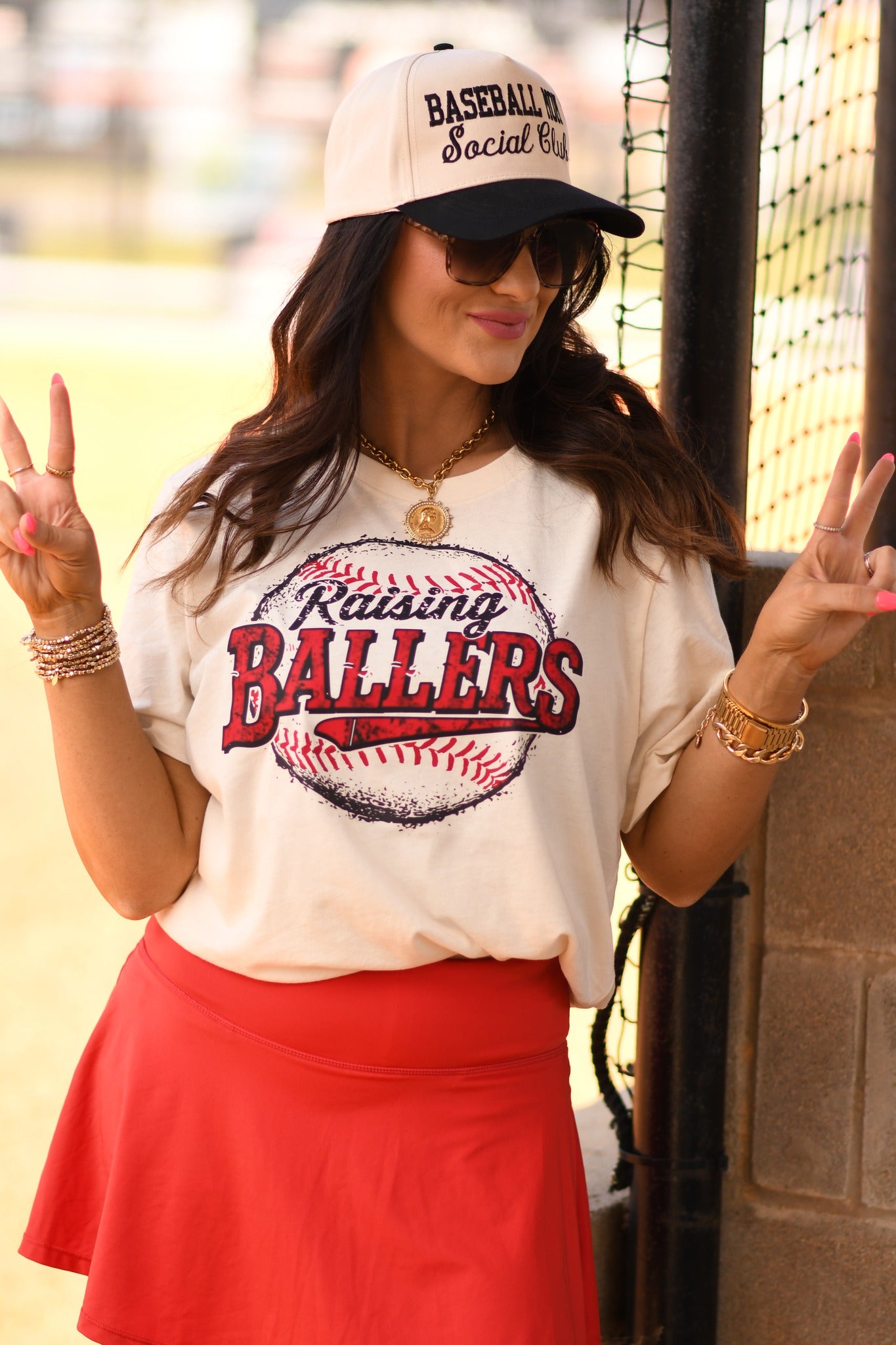 Baseball mom wearing Raising Ballers graphic tee in natural color styled with red skirt at baseball field