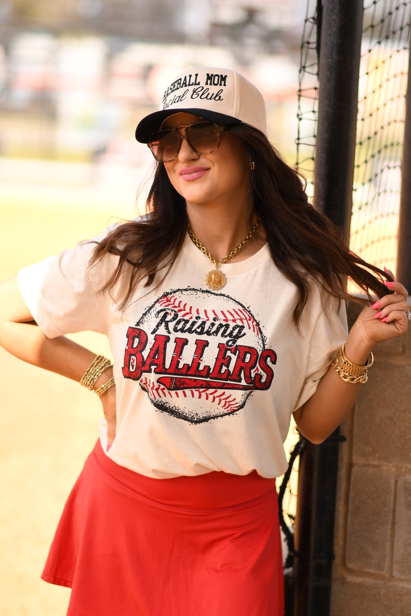 Baseball mom wearing Raising Ballers graphic tee in natural color styled with red skirt at baseball field