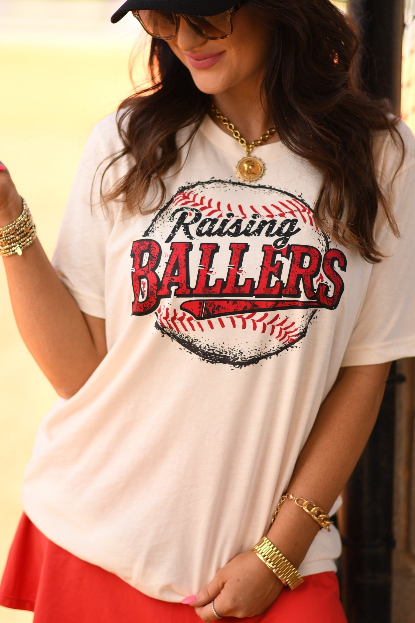 Baseball mom wearing Raising Ballers graphic tee in natural color styled with red skirt at baseball field