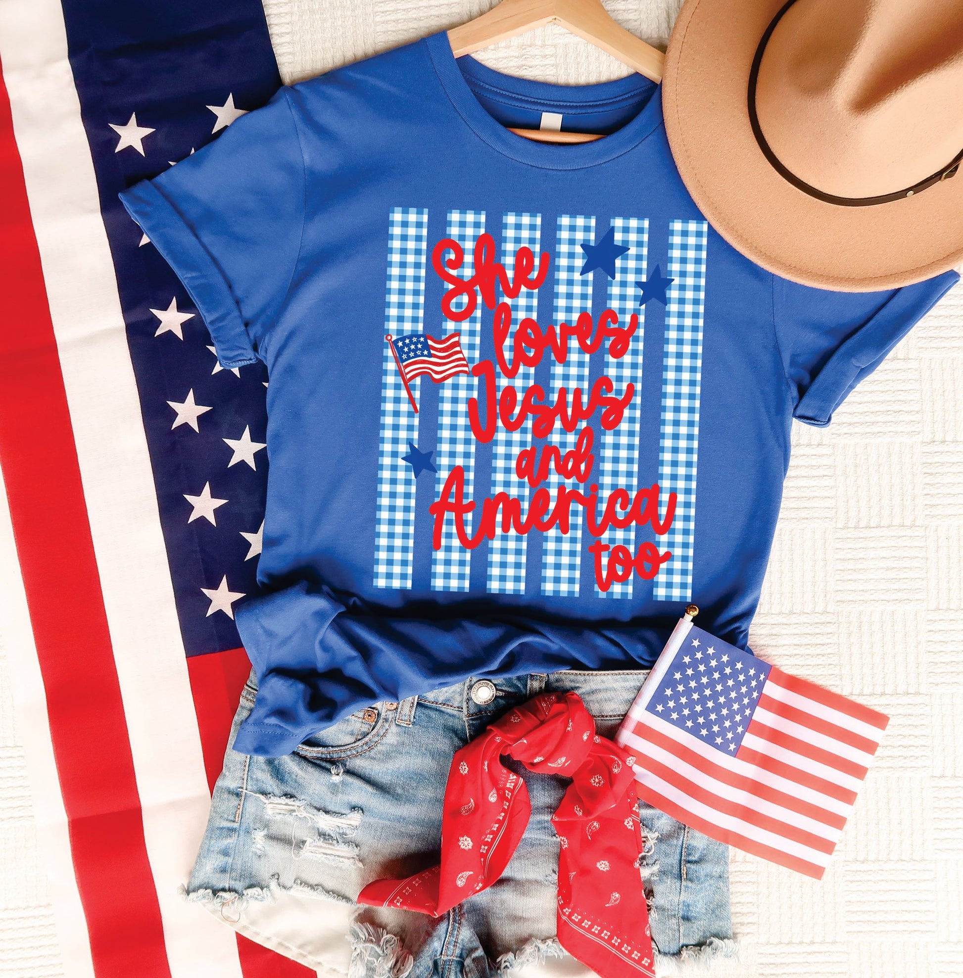 Blue She Loves Jesus and America Too patriotic graphic tee styled with denim shorts, American flag, red bandana, and tan hat.