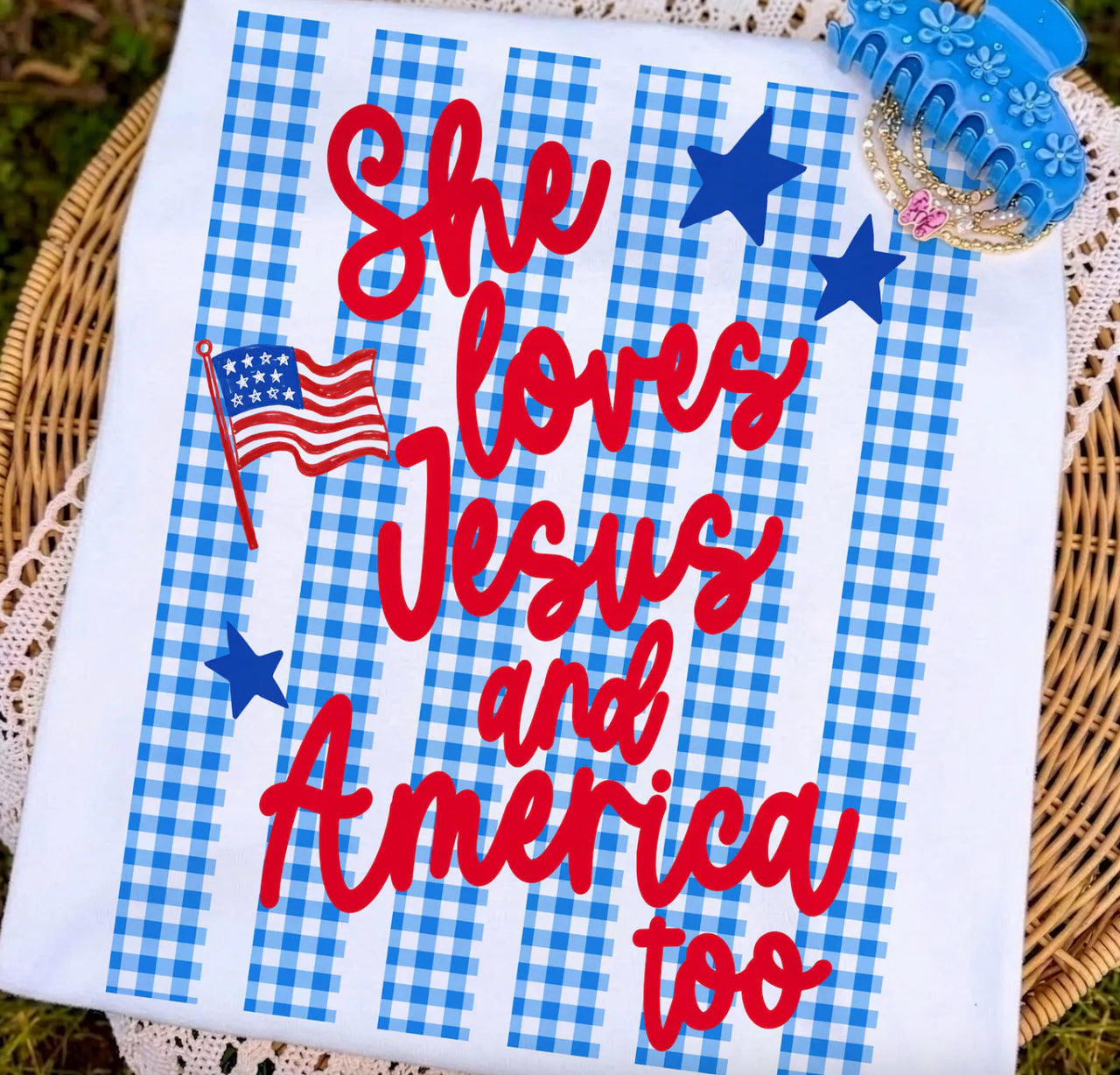 White shirt with blue gingham vertical stripes, red “She loves Jesus and America too” lettering, blue stars, and an American flag graphic.
