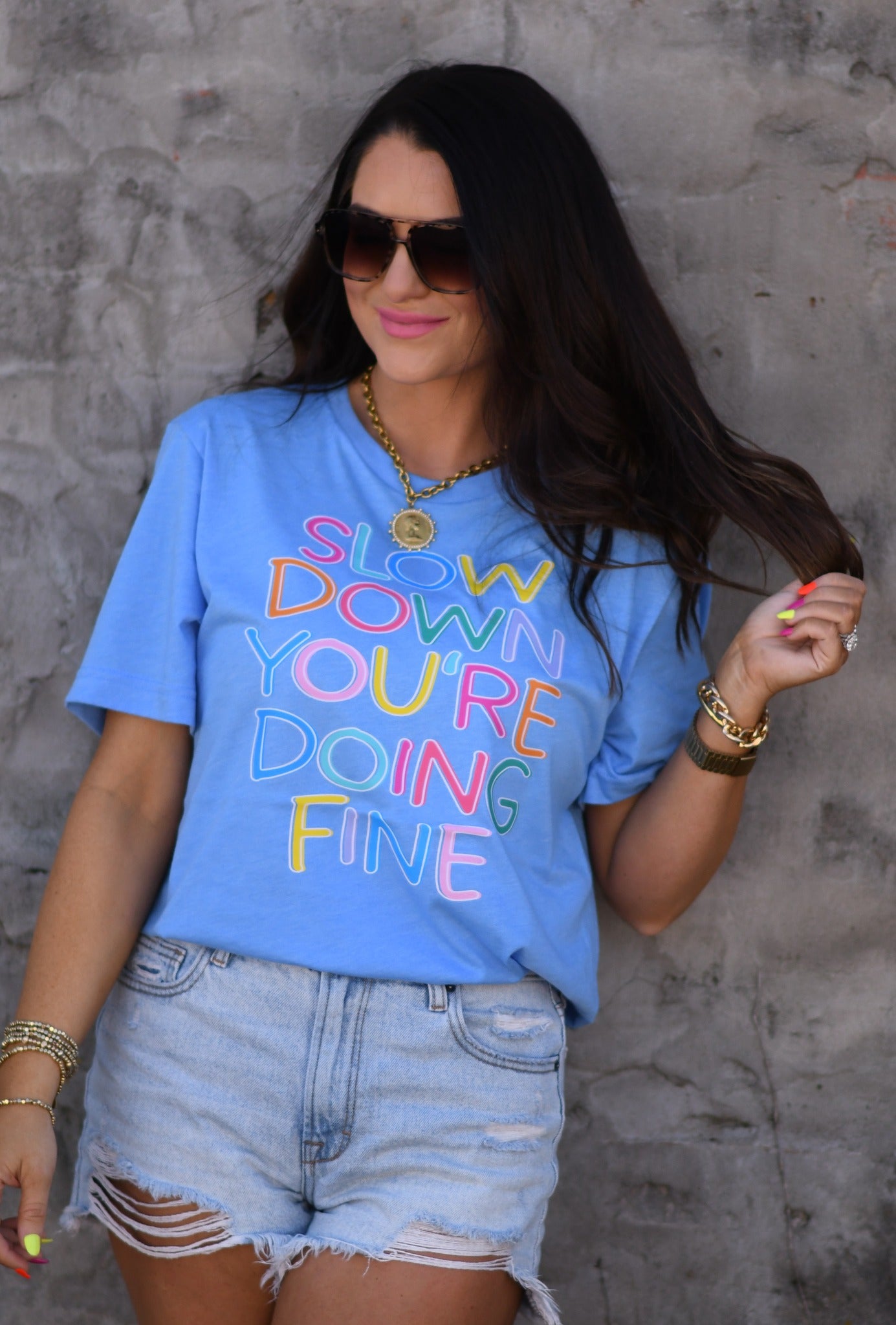Woman wearing a heather Columbia blue graphic tee with colorful “Slow Down You’re Doing Fine” text paired with distressed denim shorts and sunglasses.