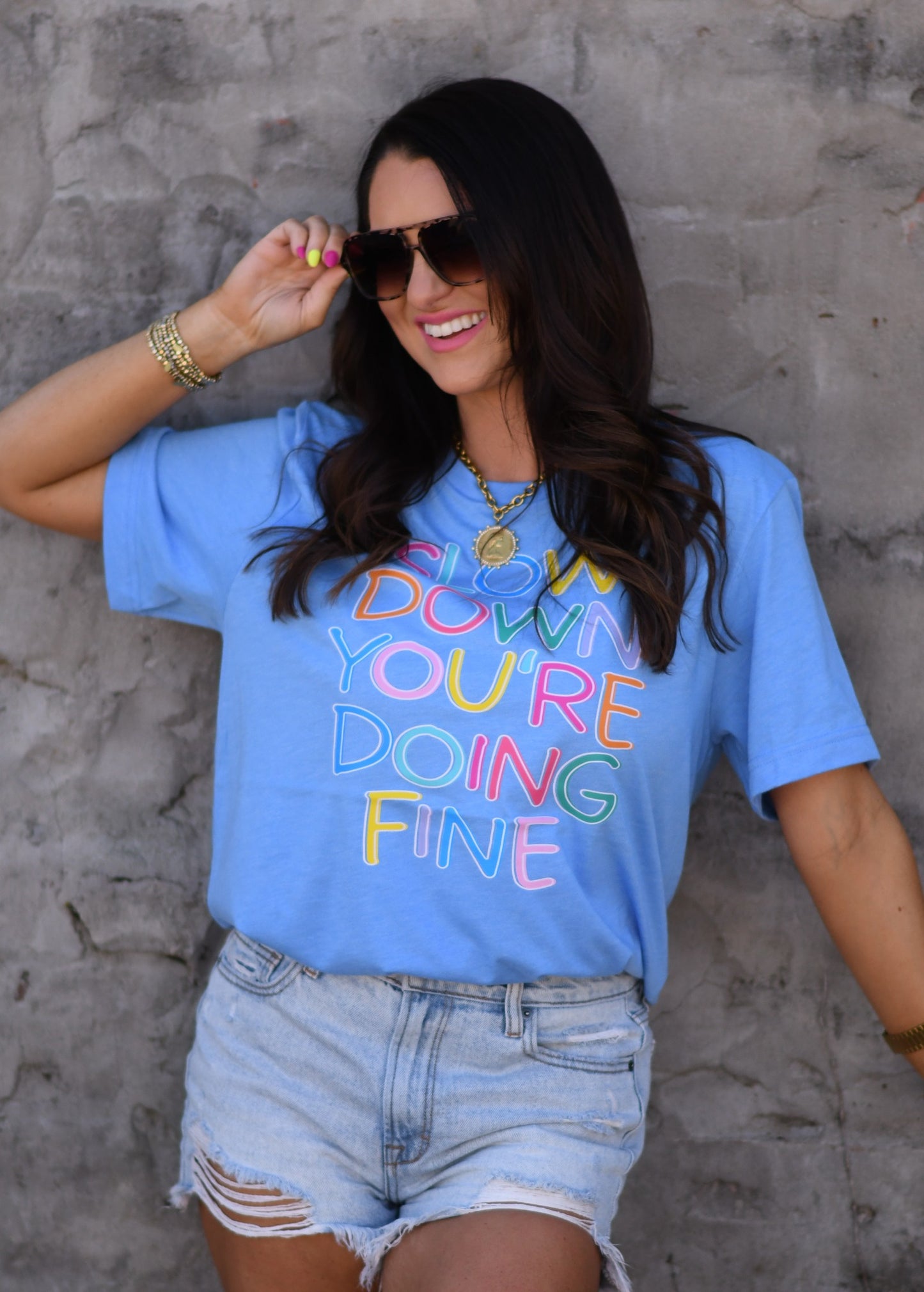 Woman wearing a heather Columbia blue graphic tee with colorful “Slow Down You’re Doing Fine” text paired with distressed denim shorts and sunglasses.