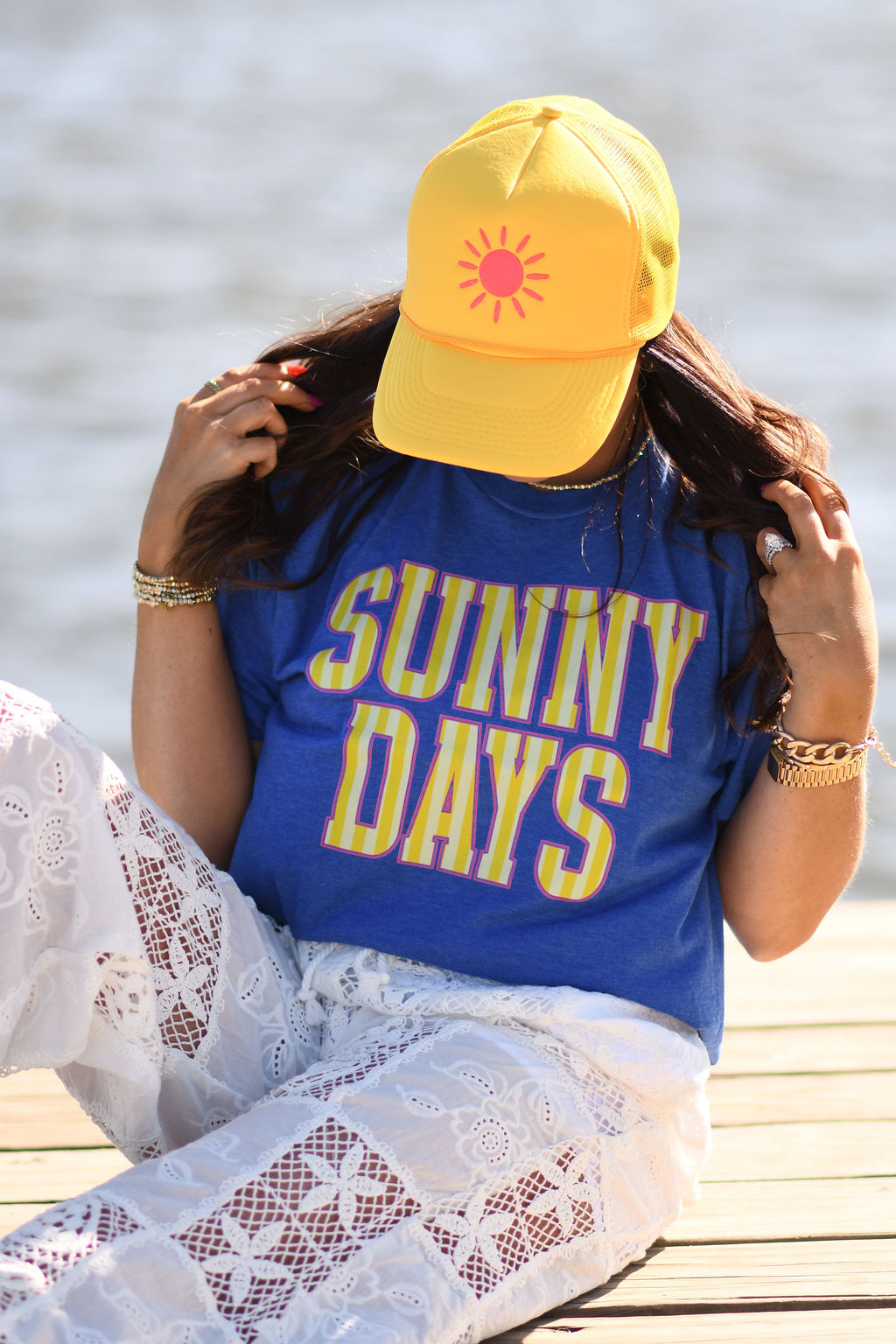 Woman wearing royal blue Sunny Days graphic tee with yellow sun trucker hat sitting outdoors