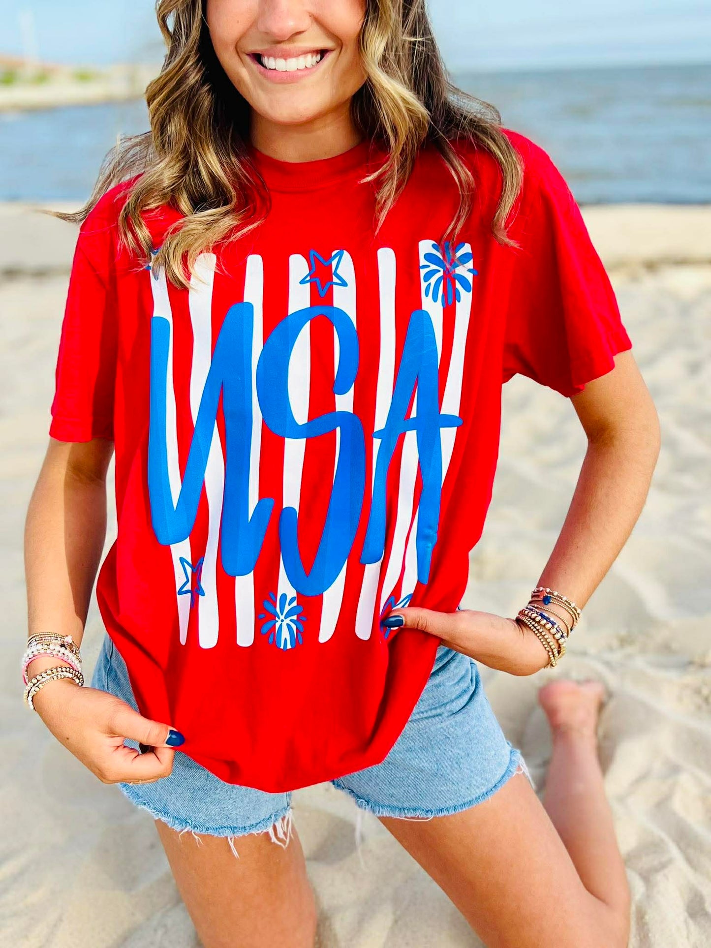 Red USA patriotic graphic t-shirt with blue lettering and white stripes worn with denim shorts on beach