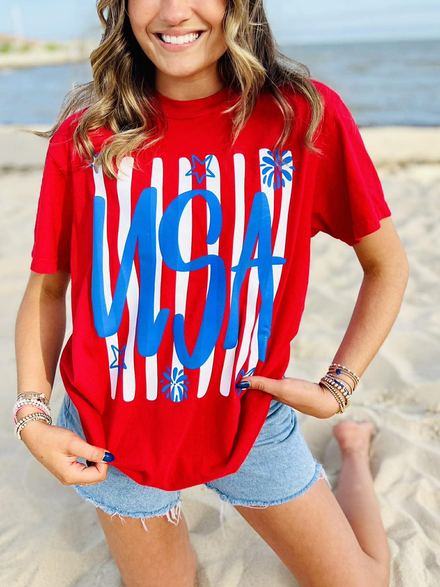 Red USA patriotic graphic t-shirt with blue lettering and white stripes worn with denim shorts on beach