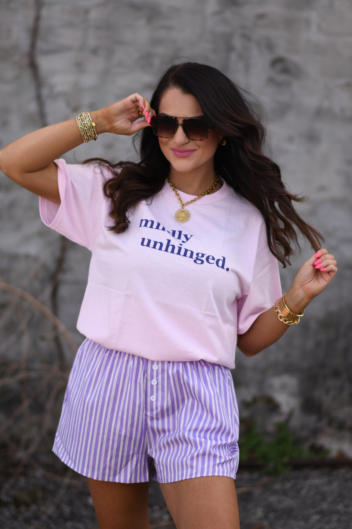 Woman wearing baby pink “mildly unhinged” graphic t-shirt with sunglasses and casual shorts, styled with gold jewelry outdoors