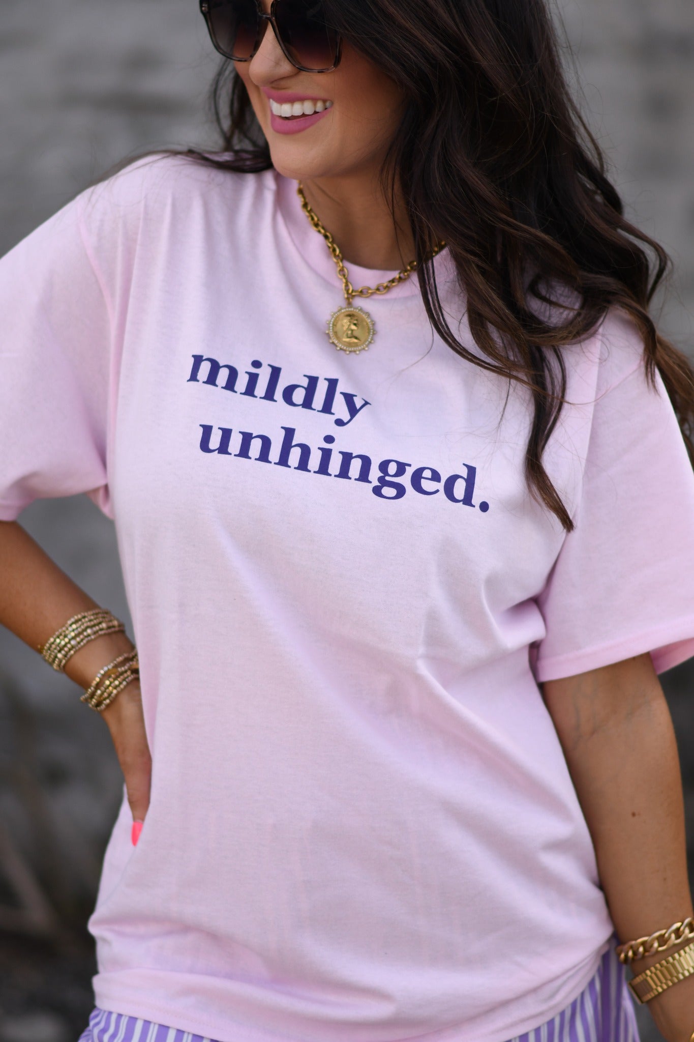 Woman wearing baby pink “mildly unhinged” graphic t-shirt with sunglasses and casual shorts, styled with gold jewelry outdoors