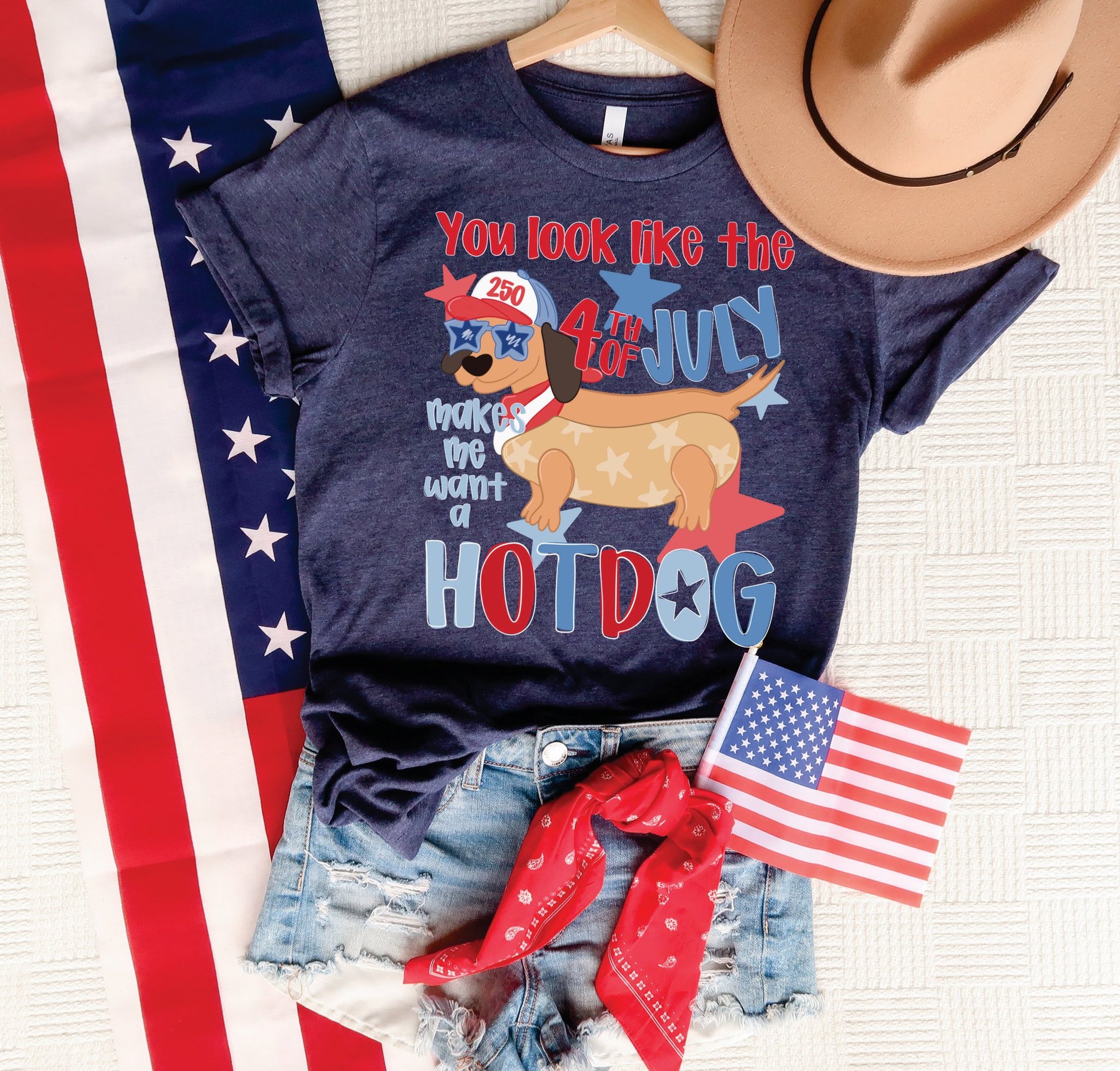 Navy patriotic 4th of July hotdog graphic tee styled with denim shorts, American flag, red bandana, and tan hat.