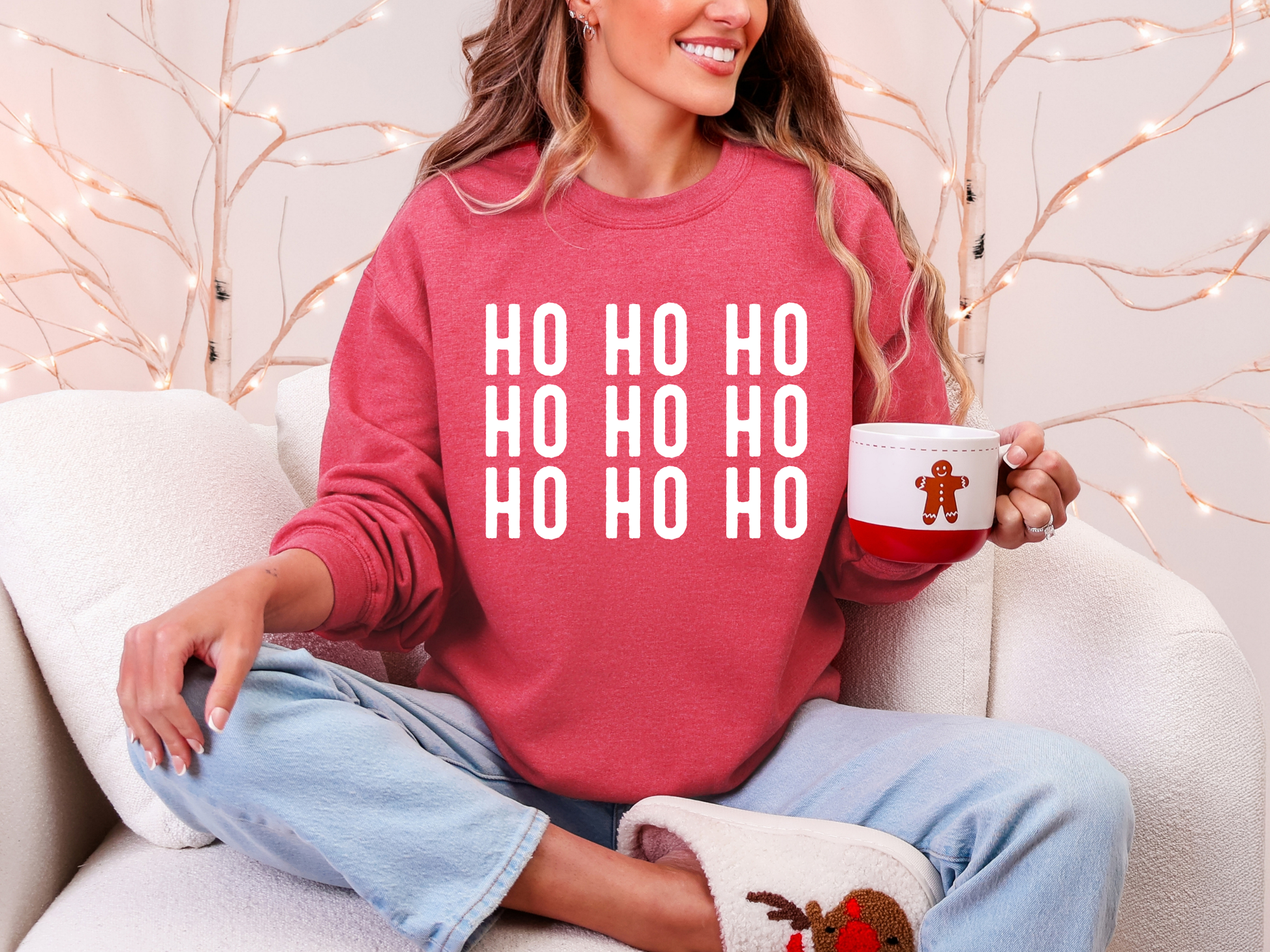 Person wearing a red sweater with 'HO HO HO' text, holding a mug, sitting on a couch with decorative lights in the background.